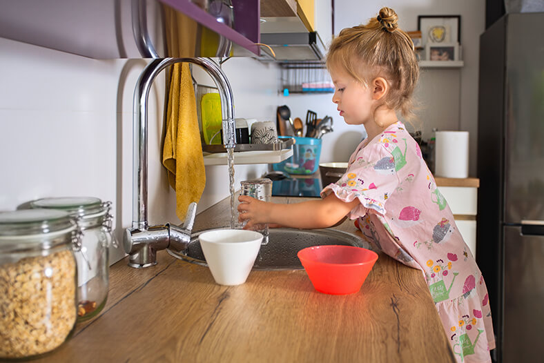 Ein Mädchen im Grundschulalter füllt ein Glas Wasser an einem laufenden Wasserhahn in der Küche.