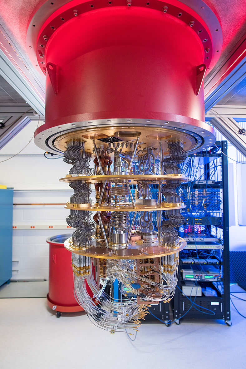 Das Bild zeigt den Quantencomputer von Google, Man sieht einen hängenden roten Zylinder, aus dem nach unten viele gebogene Leitungen führen, segmentiert von mehreren runden Scheiben. Im Hintergrund stehen Regale mit weiteren elektronischen Geräten