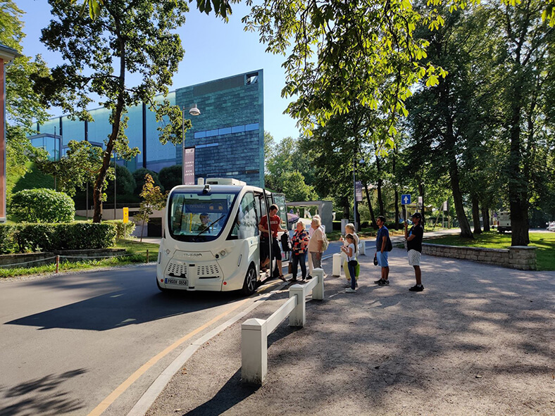 Ein weißer Bus ohne Fahrer steht auf einer Straße in Tallinn (Estland); ein junger Mann in kurzer Hose und T-Shirt steigt aus. Rechts steht eine Schlange von mehreren Menschen, die einsteigen wollen.
