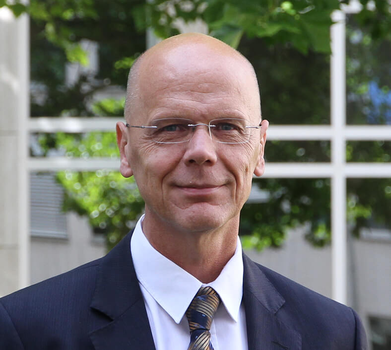 Porträt von Dr. Hüdepohl. Er hat eine Halbglatze, trägt eine Brille und einen dunklen Anzug mit Krawatte und weißem Hemd. Er steht im Freien, im Hintergrund sieht man die Fenster eines Gebäudes.