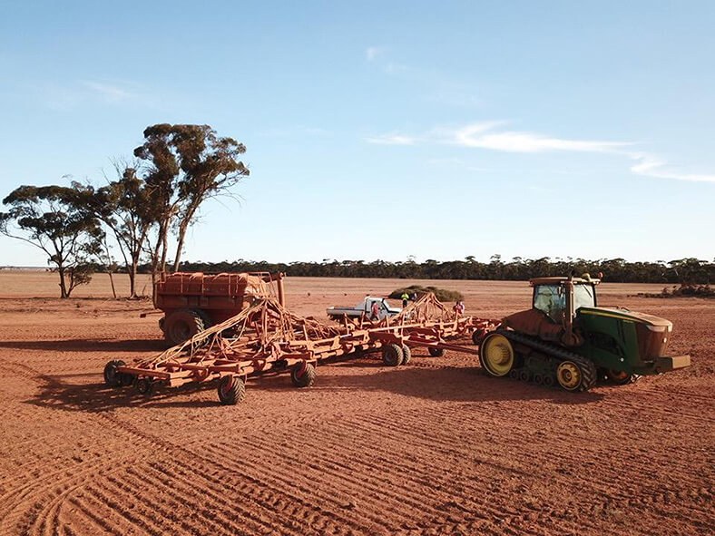 Das Bild zeigt einen Acker mit roter Erde in Australien, darauf ein Traktor und weitere Landmaschinen. Im Hintergrund ist ein Waldrand zu sehen.