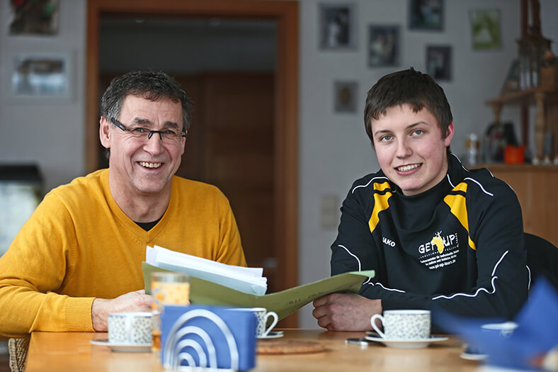 Am einem Tisch sitzen Stefan Mayr (links) und Martin Welte (rechts) vor Kaffetassen. Stefan Mayr hat eine Brille und trägt einen gelben Pullover, Martin Welte trägt eine schwarz-gelbe Trainingsjacke. Sie blättern zusammen in Unterlagen.