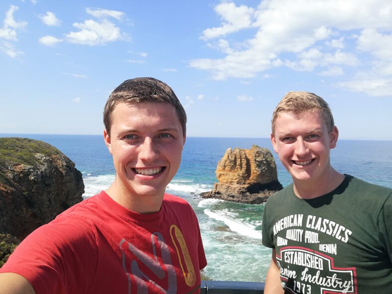 Martin Welte (links) trägt ein rotes T-Shirt, sein Bruder steht rechts neben ihm hat blonde Haare und trägt ein grünes T-Shirt. Im Hintergrund sieht man Meer, Felsen und blauen Himmel.