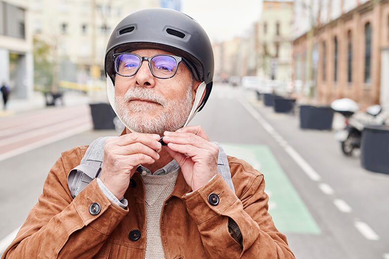 Alterer Mann mit birlle und Vollbart steht auf der Straße und befestigt einen Fahrradhelm auf seinem Kopf.