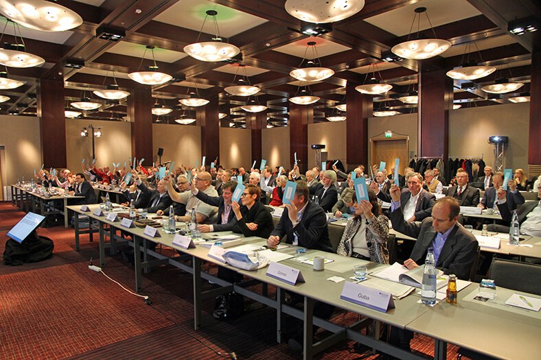 Blick auf die Teilnehmer der Vertreterversammlung der BG ETEM in einem Saal in Köln. Sie sitzen an mehreren langen Tischreihen und strecken für eine Abstimmung blaue Zettel in die Höhe.