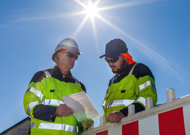 Zwei Arbeiter mit Schutzkleidung, Kopfbedeckung und Sonnenbrille stehen im Freien im Sonnenschein und sehen zusammen auf ein Dokument.