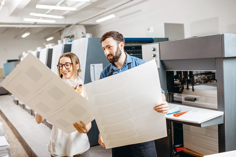 Nach einem schweren Jahr 2020 blickt die Druckindustrie wieder positiver in die Zukunft. Mann und junge Frau mit Brille stehen in einer Produktionshalle einer Druckerei und halten große Druckbögen in den Händen, auf die sie lächelnd schauen.