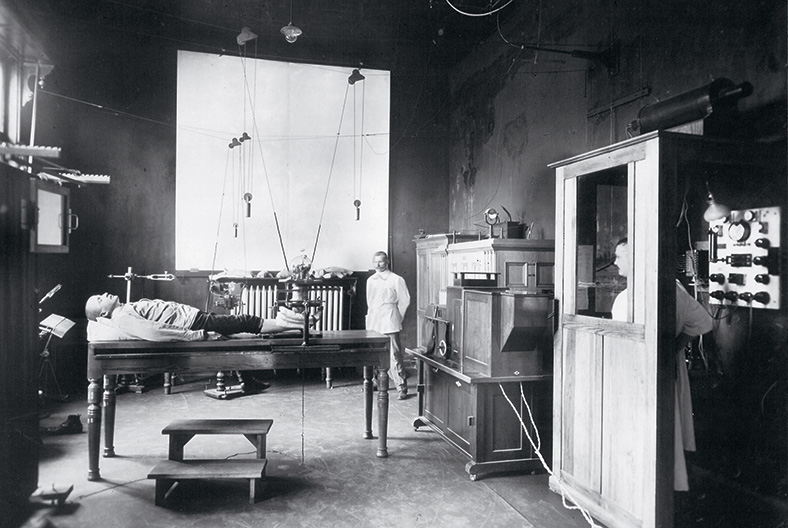 Schwarzweißaufnahme eines Benhandlungsraums, auf einer Liege liegt ein Mann und wird mit einem historischen Gerät geröntgt, ein Arzt steht seitlich im Hintergrund, ein anderer Mitarbeiter bedient das Röntgengerät aus einer Kammer heraus.