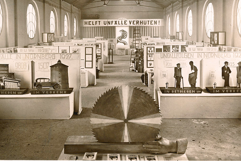 Schwarzweiß-Aufnahme eines Ausstellungsraums mit einem großen Kreissägenblatt im Vordergrund, dahinter rechts und links Stellwände mit Bildern von Unfallfolgen.