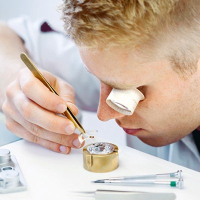 Ein junger Mann hat ein Vergrößerungsglas am Auge und arbeitet mit einer Pinzette an einer Uhr.
