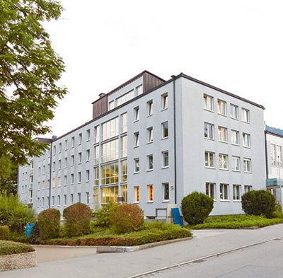 Ansicht der BG Klinik Bad Reichenhall. Ein weißes, mehrstöckiges Gebäude mit vielen Fenstern und Flachdach, umgeben von Büschen.