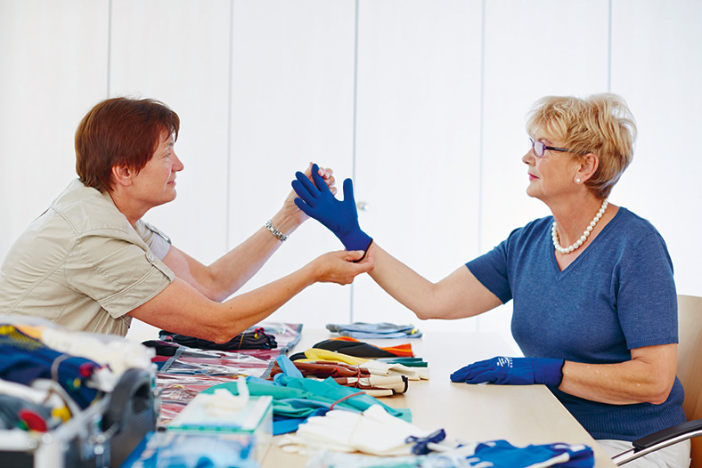Schutzhandschuhberatung in der BG Klinik Bad Reichenhall. Eine Mitarbeiterin testet an einer Patientin einen blauen Handschuh. Beide sitzen sich gegenüber an einem Tisch voller bunter Handschuhe.