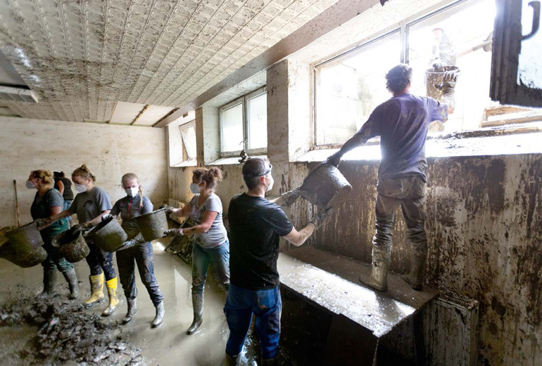Eine Menschenkette mit Mund-Nasen-Masken im Inneren eines durchnässten Untergeschoss-Raums mit lehmfarbenem Wasser und Steinen auf dem Boden. Die Menschenkette reicht sich Eimer weiter, die schließlich durch ein Fenster nach draußen gelangen.