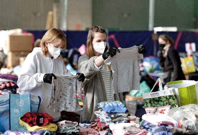 Ein Hallen-Flohmarkt, auf dem zwei Frauen mit Handschuhen und Mund-Nasen-Masken vor einem Stand mit Kleidung stehen und jeweils ein T-Shirts hochhalten und begutachten.  Sorgsamer Umgang mit Geld ist selbstvers