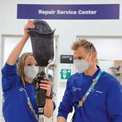 Eine Frau und ein Mann in Blau gekleidet und mit Mund-Nasen-Masken betrachten eine Beinprothese. Dahinter im Raum steht auf einem Schild „Repair Service Center“.