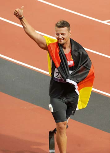 Ein Mann in Sportlerkleidung mit Teilnehmernummer auf dem T-Shirt geht ein Sprinterfeld entlang. Er macht das Daumen-hoch-Zeichen, hat eine Deutschlandfahne umgehängt und trägt links eine Beinprothese.