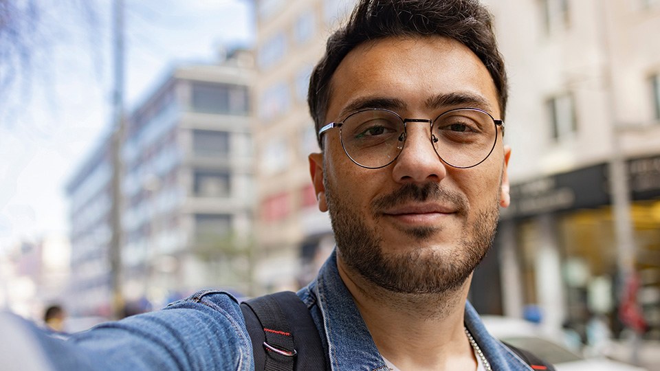 Junger bärtiger Mann mit Brille schaut bei einem Selfie in die Kamera, im Hintergrund Stadtbild