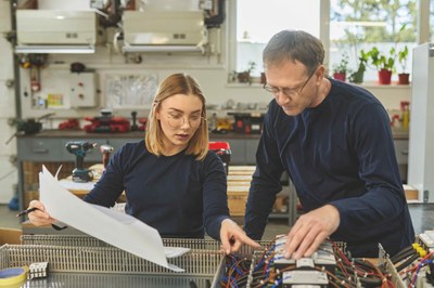 Ein Mann und eine Frau  vergleichen ein Bauteil mit einem Bauplan. 