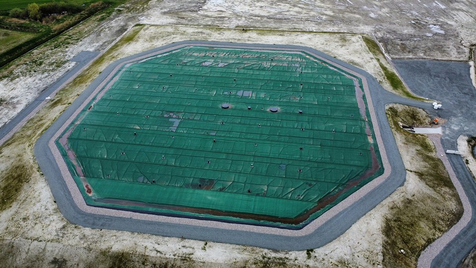 Luftaufnahme eines großflächigen, mit grüner Plane abgedeckten Erdbeckenspeichers für Fernwärme in Schleswig-Holstein